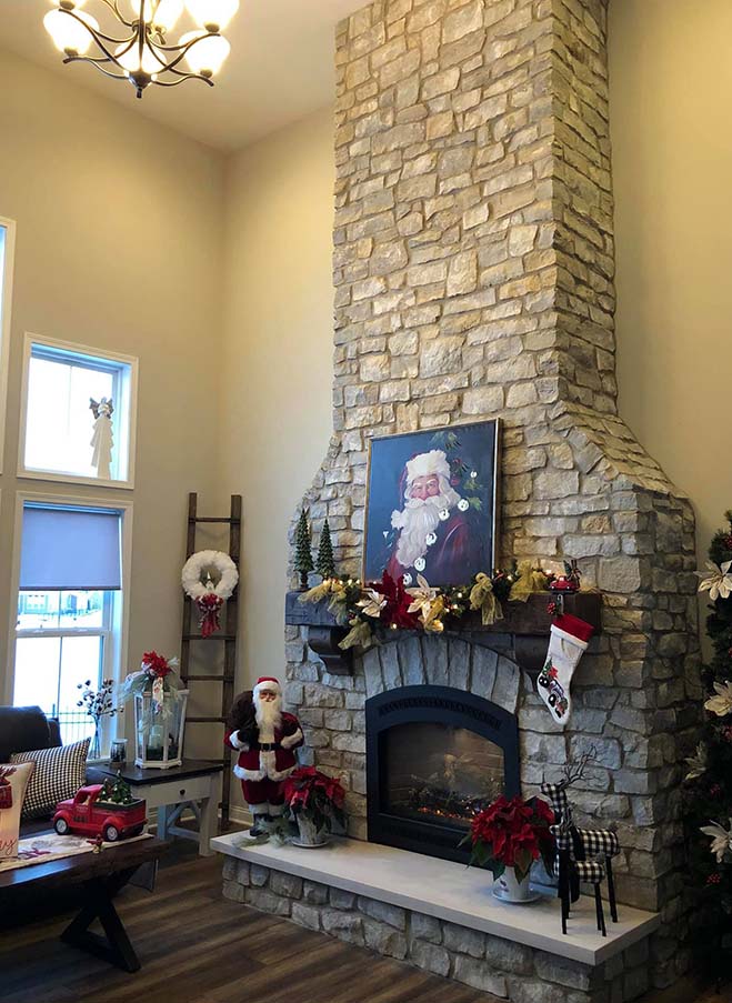 A gray stone fireplace decorated for Christmas with garland and a stocking. There is a miniature Santa and a painting of santa on the mantel.