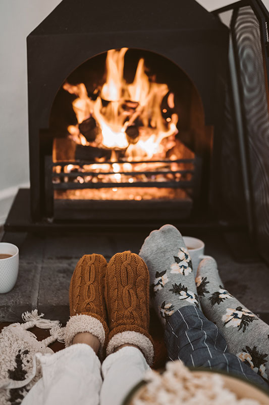 Two people's feet cuddled up in front of the fireplace. The people are wearing socks and long pjs. The have coffee mugs resting on the hearth