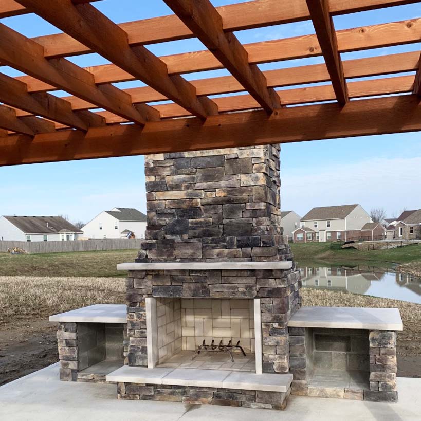 A stacked stone outdoor fireplace with houses in the background and a wood awning in the foreground with a blue sky.