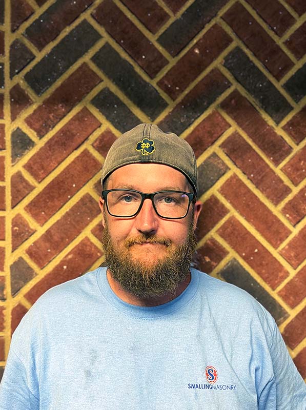 Carl Tanner - Lead Technician - Standing in front of a brick wall wearing a brown hat and a blue Smalling T.  He is wearing glasses, has a light brown mustache and beard.