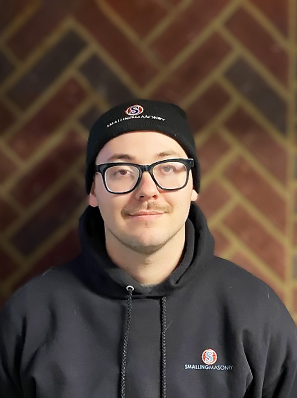 Jacob Ondeka - Consultant - Standing in front of brick wall wearing a black stocking hat and black hoodie with Smalling Logo.  He is wearing black rimmed glasses with a mustache and nice smile.
