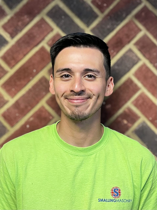 Jorge Doren - Lead Technician - Dark hair and with with mustache and goatee with some dimples.  He is wearing a green Smalling T.  Standing in front of a brick wall.