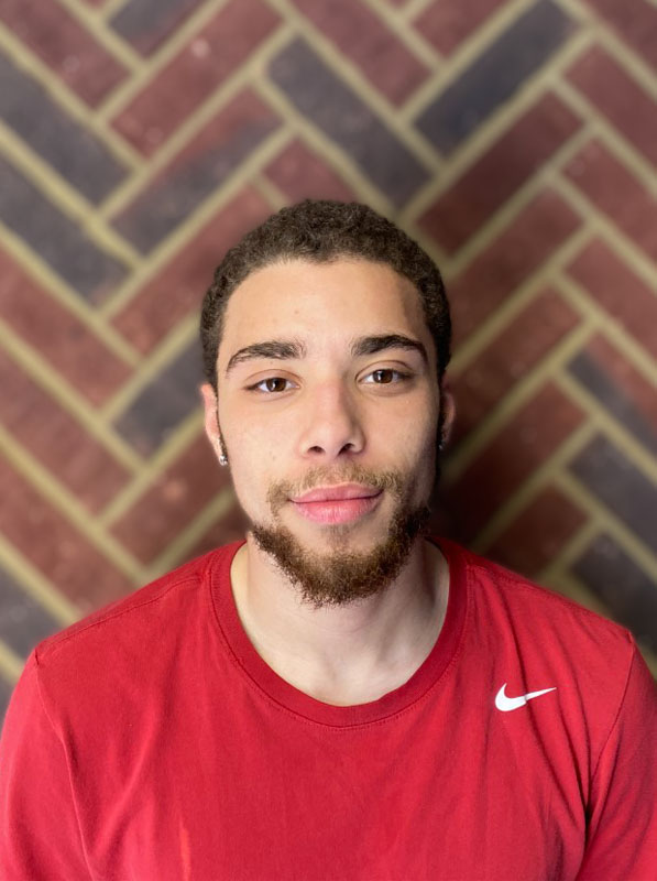 Jaylan Perkins - Apprentice - He is standing in front of a brick wall.  He has dark brown short hair, mustache and beard.  He has a very pleasant smile on his face.   He is wearing a red Nike T.