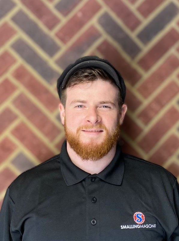 Roger Thompson - Consultant - Standing in front of a brick wall with a black hat and T with Smalling logo.  Short reddish hair, mustache and beard with a pleasant face.
