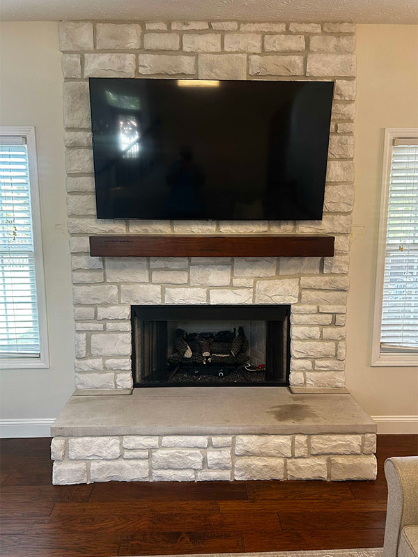 A new white stone fireplace with a wooden mantelpiece after remodel.  A tv hangs over the mantel.