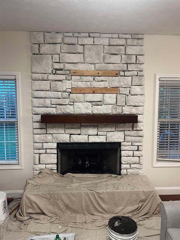 A white stone breast wall and windows on each side with a thick wooden mantel during a remodel with the floor and hearth covered with protective drop clothes.