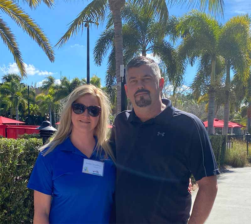 Gary and Jenny (owners) standing in front of palm trees. She has long blonde hair wearing sunglasses and blue buttondown and he has a mustache and goatee with close shaven hair and a black buttondown.