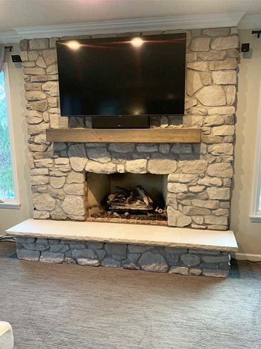 Gray rock fireplace with a light brown wood mantelpiece and granite hearth with TV mounted above mantel and windows on each side.