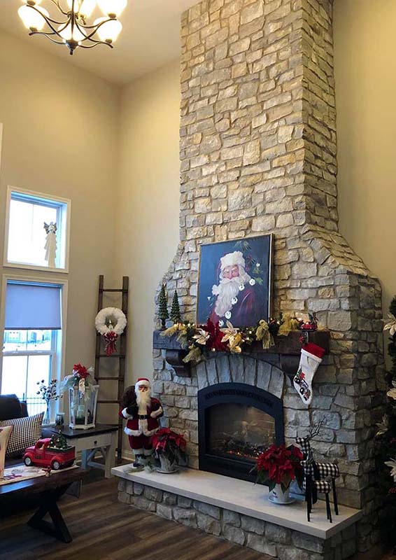 A gray stone fireplace decorated for Christmas with garland and a stocking. There is a miniature Santa and a painting of santa on the mantel.