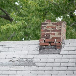 a damaged and crumbling masonry chimney on a gray roof with trees in the background