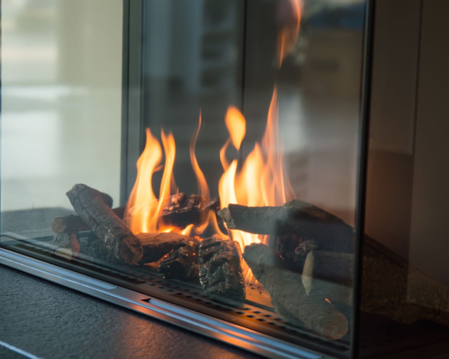 stock image shows a modern gas fireplace with a glass enclosure, featuring realistic ceramic logs arranged over a metal grate. Bright orange flames rise between the logs, creating a warm and cozy atmosphere. The sleek glass panels and dark stone or metal base give the fireplace a contemporary and polished look, allowing the fire to be the main focal point.